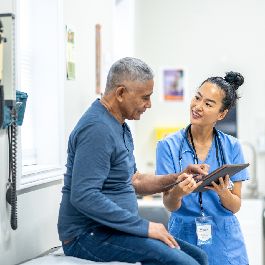 A senior gentleman of Hispanic decent, sits up on an exam table during a routine check-up at the doctors. A nurse, who is wearing blue scrubs, is standing in front of the gentleman as she reviews some recent test results with the patient on a tablet.