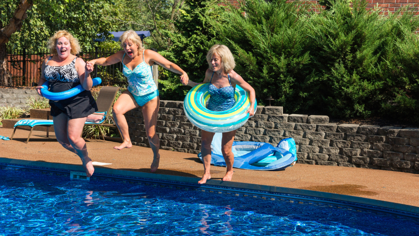 Three mature women jumping into a swimming pool, laughing and wearing tubes.