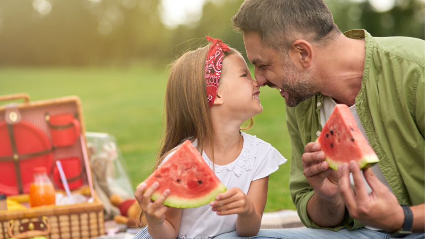 Adorable little girl playing with her loving daddy while eating watermelon, family having a picnic in the green park on a summer day.