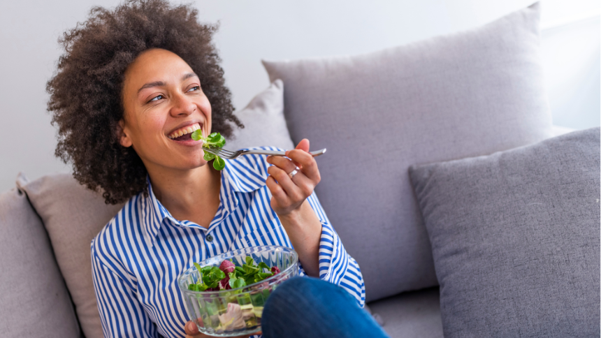 Close-up of a Black woman eating a salad in the living-room while smiling because she understands the truth behind common nutrition myths.