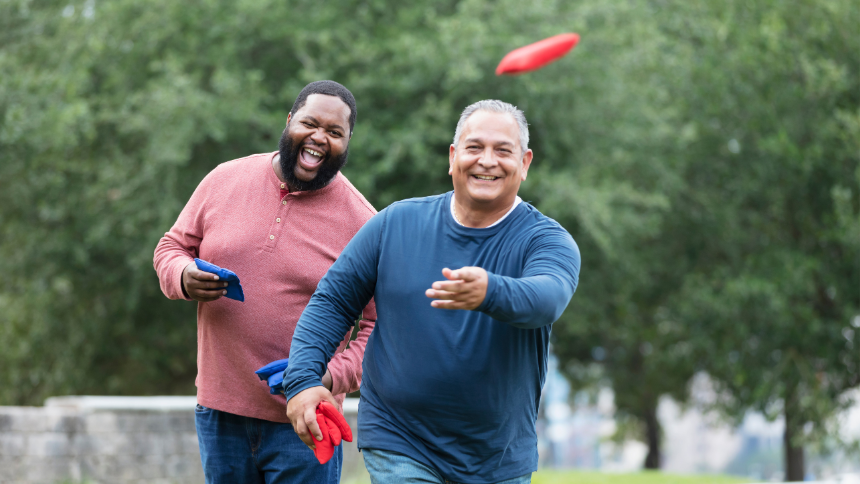 Two plus size multiracial men having fun at the park playing a bean bag toss game and smiling because they are managing diverticulosis.