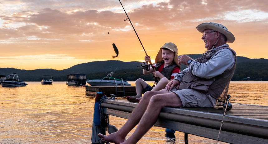 A grandfather is teaching his grandson to fish in summer. They are both on the dock/pier and laughing. It is a beautiful summer day. Across the lake, there is a mountain. The little boy just caught a fish and is very proud. The sky is multicolored during this beautiful sunset. 