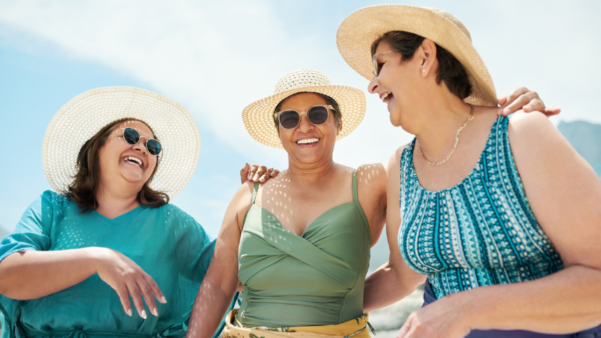 A mature group of friends standing together during a day out on the beach. They are wearing hats and sunglasses to protect their skin from cancer.