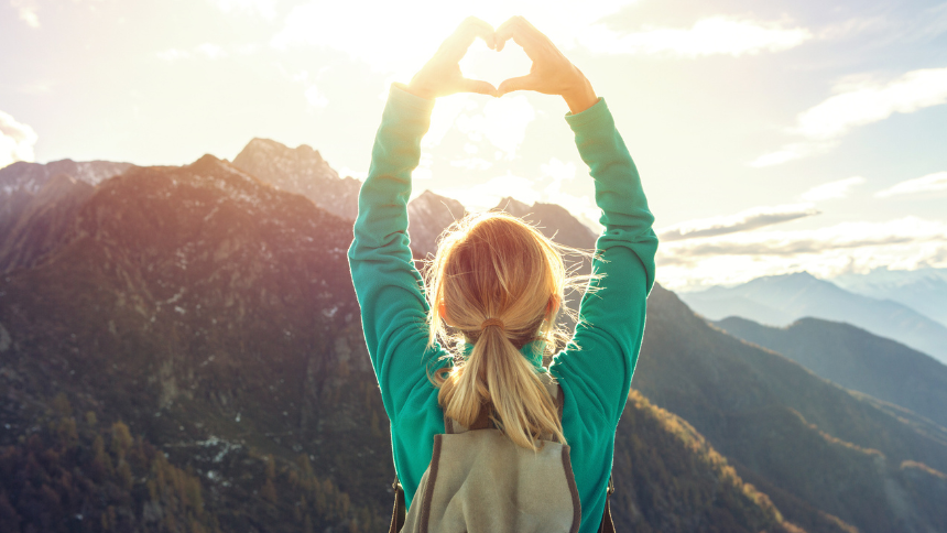 A woman with blond hair wearing a green long-sleeve shirt and backpack overlooking the mountains and making a heart shape with her hands.